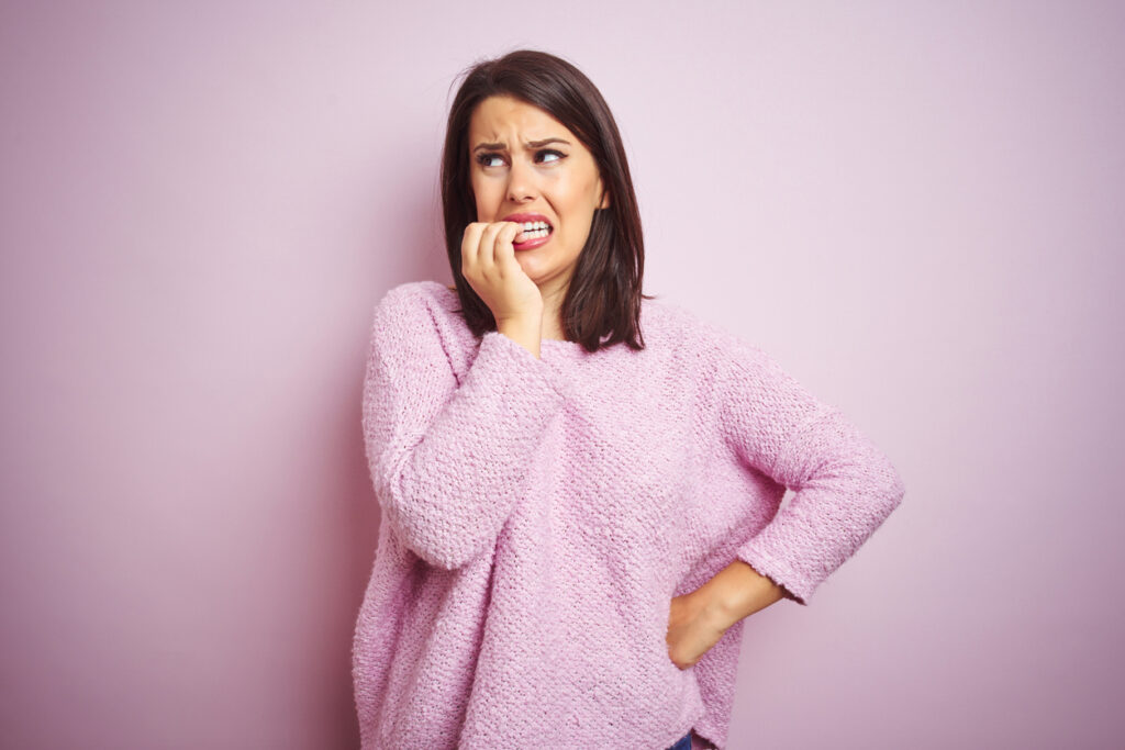 young beautiful brunette woman wearing a sweater over pink isolated background looking stressed and nervous with hands on mouth biting nails. anxiety problem.